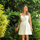 Mujer frente a un jardin luciendo un vestido blanco corto con detalles de botones y manguillos finos