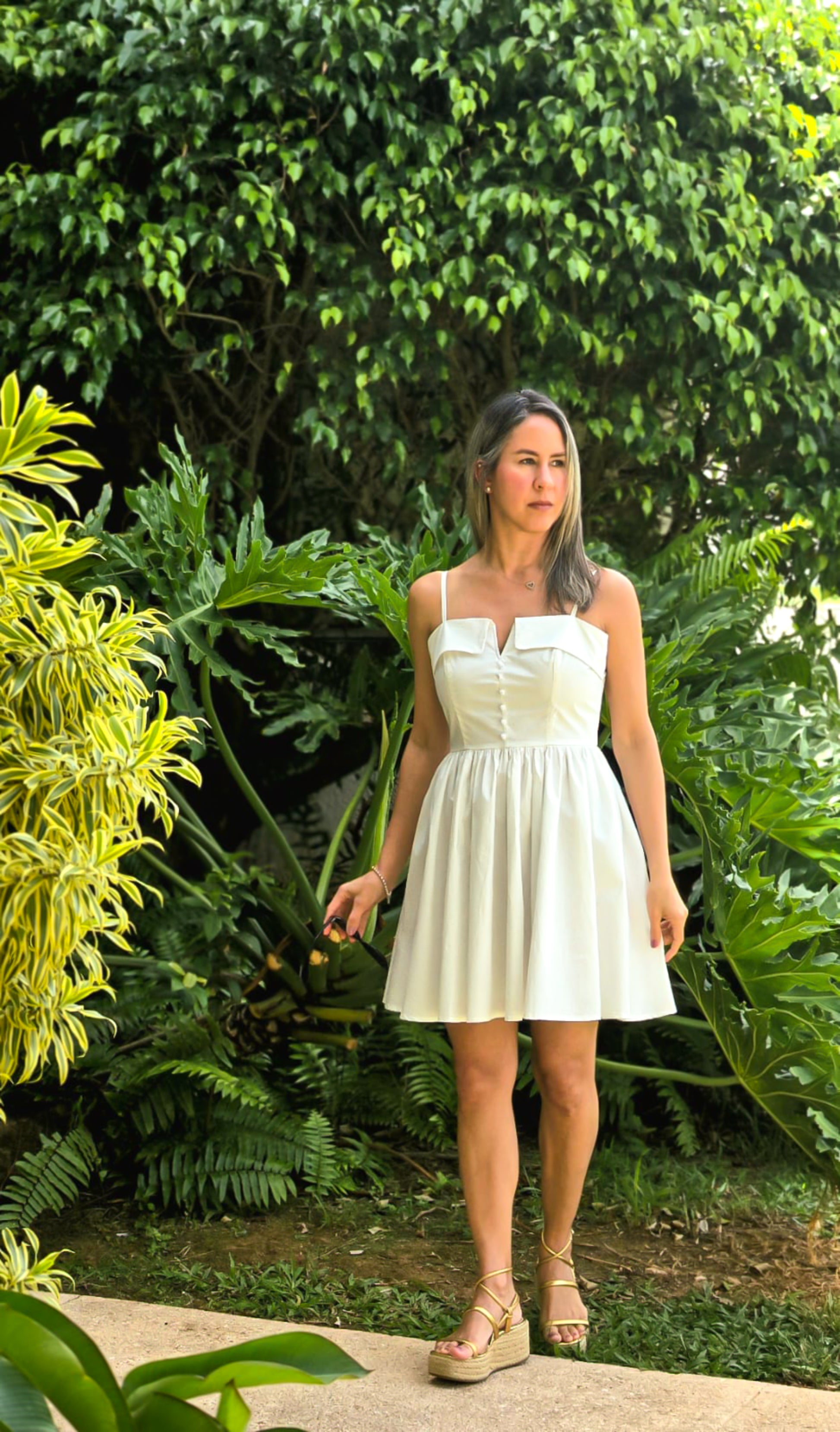 Mujer frente a un jardin luciendo un vestido blanco corto con detalles de botones y manguillos finos