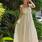 "Mujer usando un vestido blanco largo con tirantes delgados y detalle de lazo en el escote, ideal para verano. Fondo de jardiens naturales. Look fresco, elegante y minimalista."