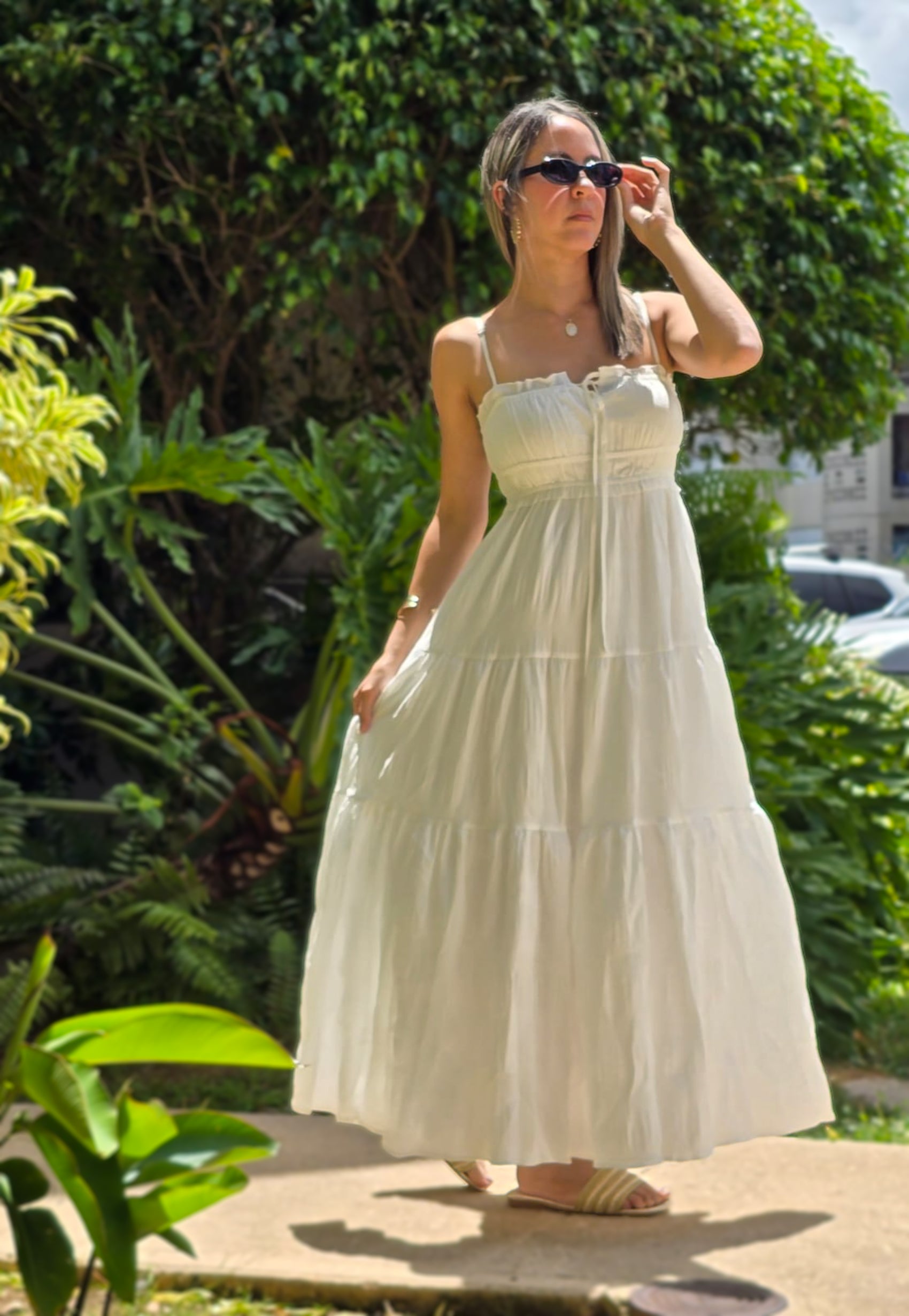 "Mujer usando un vestido blanco largo con tirantes delgados y detalle de lazo en el escote, ideal para verano. Fondo de jardiens naturales. Look fresco, elegante y minimalista."