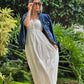 "Mujer usando un vestido blanco largo con tirantes delgados y detalle de lazo en el escote y una chaqueta de mahon sobrepuesta, ideal para un look casual. Fondo de jardiens naturales. Look casual elegante y minimalista."