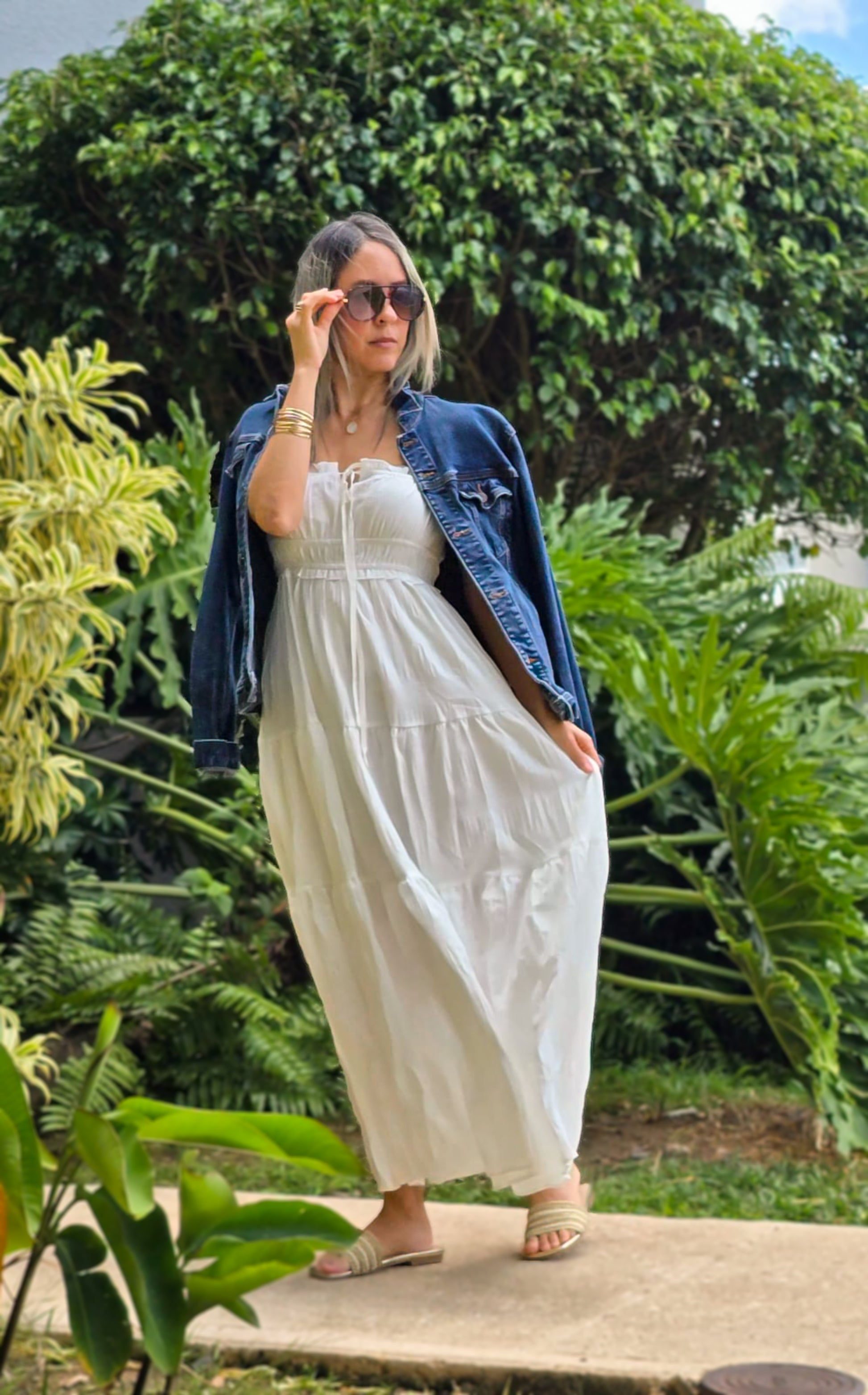 "Mujer usando un vestido blanco largo con tirantes delgados y detalle de lazo en el escote y una chaqueta de mahon sobrepuesta, ideal para un look casual. Fondo de jardiens naturales. Look casual elegante y minimalista."