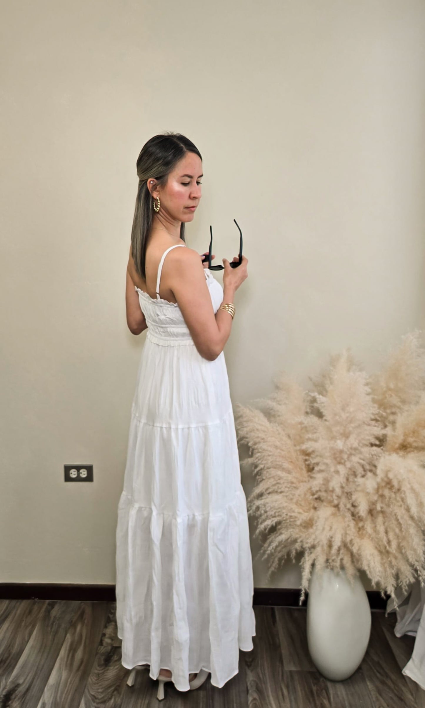 "Mujer de espalda usando un vestido blanco largo con tirantes delgados y detalle de lazo en el escote, ideal para verano. Fondo neutro con decoración natural de pampas. Look fresco, elegante y minimalista."