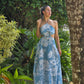 Mujer usando vestido maxi azul con estampado floral blanco, escote halter y detalles cutout frente a jardines naturales. Diseño elegante ideal para verano o vacaciones