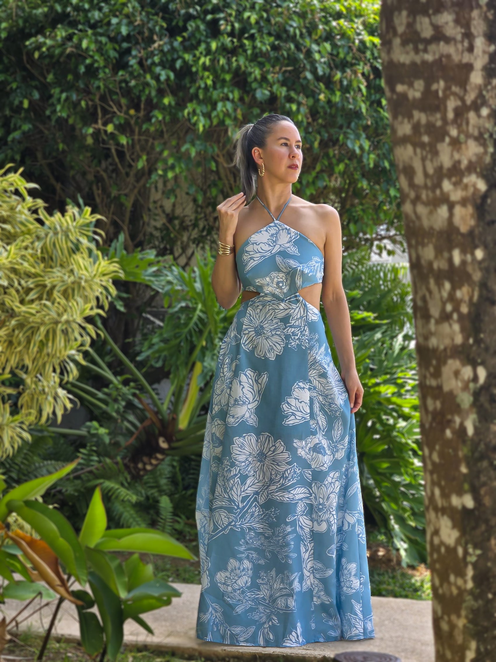 Mujer usando vestido maxi azul con estampado floral blanco, escote halter y detalles cutout frente a jardines naturales. Diseño elegante ideal para verano o vacaciones