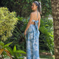 Mujer de espalda usando vestido maxi azul con estampado floral blanco, escote halter y detalles cutout frenta a jardines naturales. Diseño elegante ideal para verano o vacaciones