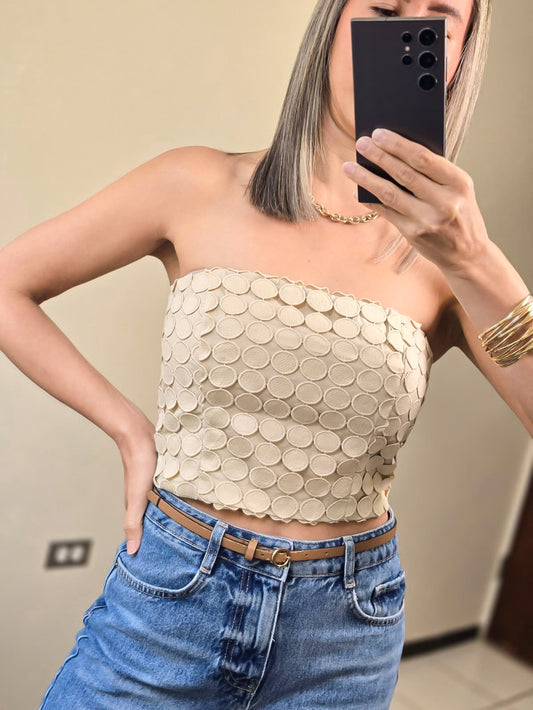 Imagen de plano medio de mujer con cabello color rubio vistiendo un statement piece (en forma de tubo) con diseño calado (circulos) en color beige tipo crop y un mahón azul high waist.  frente a un fondo neutral.