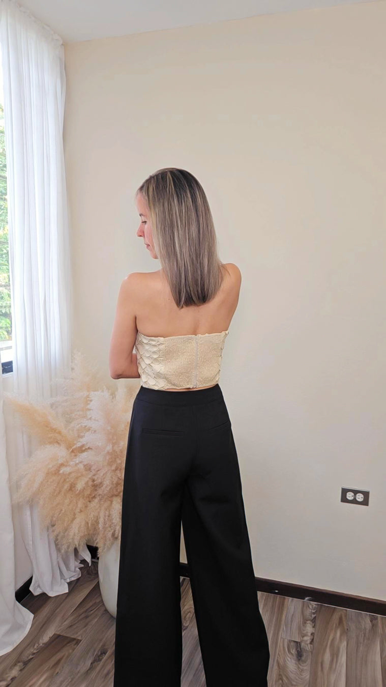 Imagen de espalda de mujer con cabello color rubio mostrando la parte trasera de un statement piece (en forma de tubo) con diseño calado (circulos) en color beige tipo crop y elasticos con cierre de zipper.   y un pantalón negro estructurado.  frente a un fondo neutral.