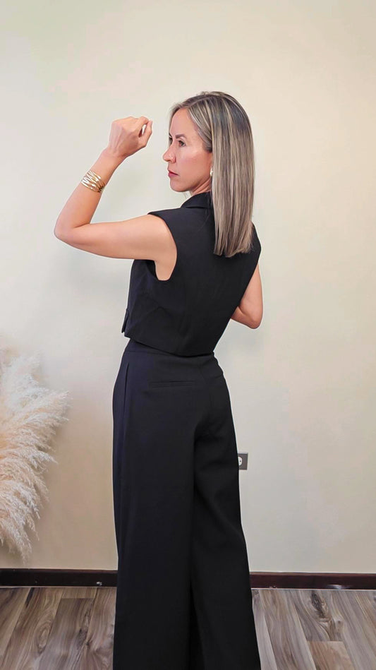 Imagen de espalda de mujer con cabello color rubio visitiendo un set de vest sin manga con botones frontales y pantalon a juego en color negro frente a un fondo neutro.  Este set es una pieza premium de la coleccion de new arrivals de la boutique Enso Amara.