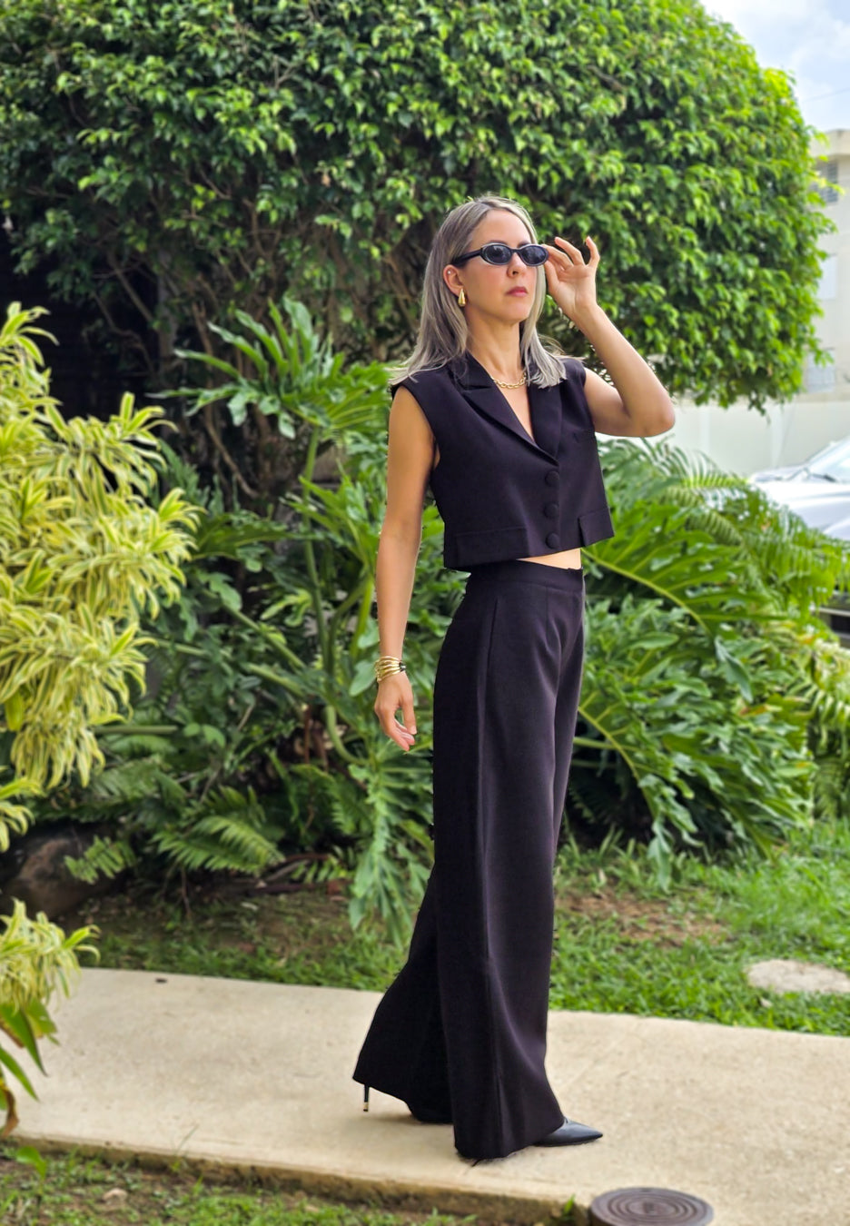 Imagen de mujer con cabello color rubio visitiendo un set de vest sin manga con botones frontales y pantalon a juego frente a un jardin llamativo con colores verdes.  Utilizando unas gafas para el sol color negras.  Este set es una pieza premium de la coleccion de new arrivals de la boutique Enso Amara.