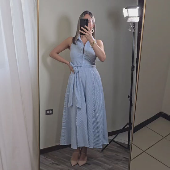 Mujer rubia vistiendo un vestido halter con cuello de camisa, botones frontales, cinta para ajustar en la cintura y una abertura en el frente de la falda.  El vestido es blanco con lineas azules verticales.
