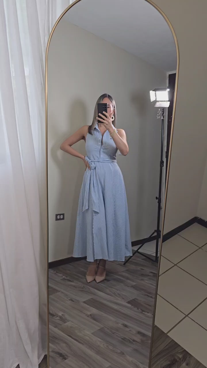 Mujer rubia vistiendo un vestido halter con cuello de camisa, botones frontales, cinta para ajustar en la cintura y una abertura en el frente de la falda.  El vestido es blanco con lineas azules verticales.